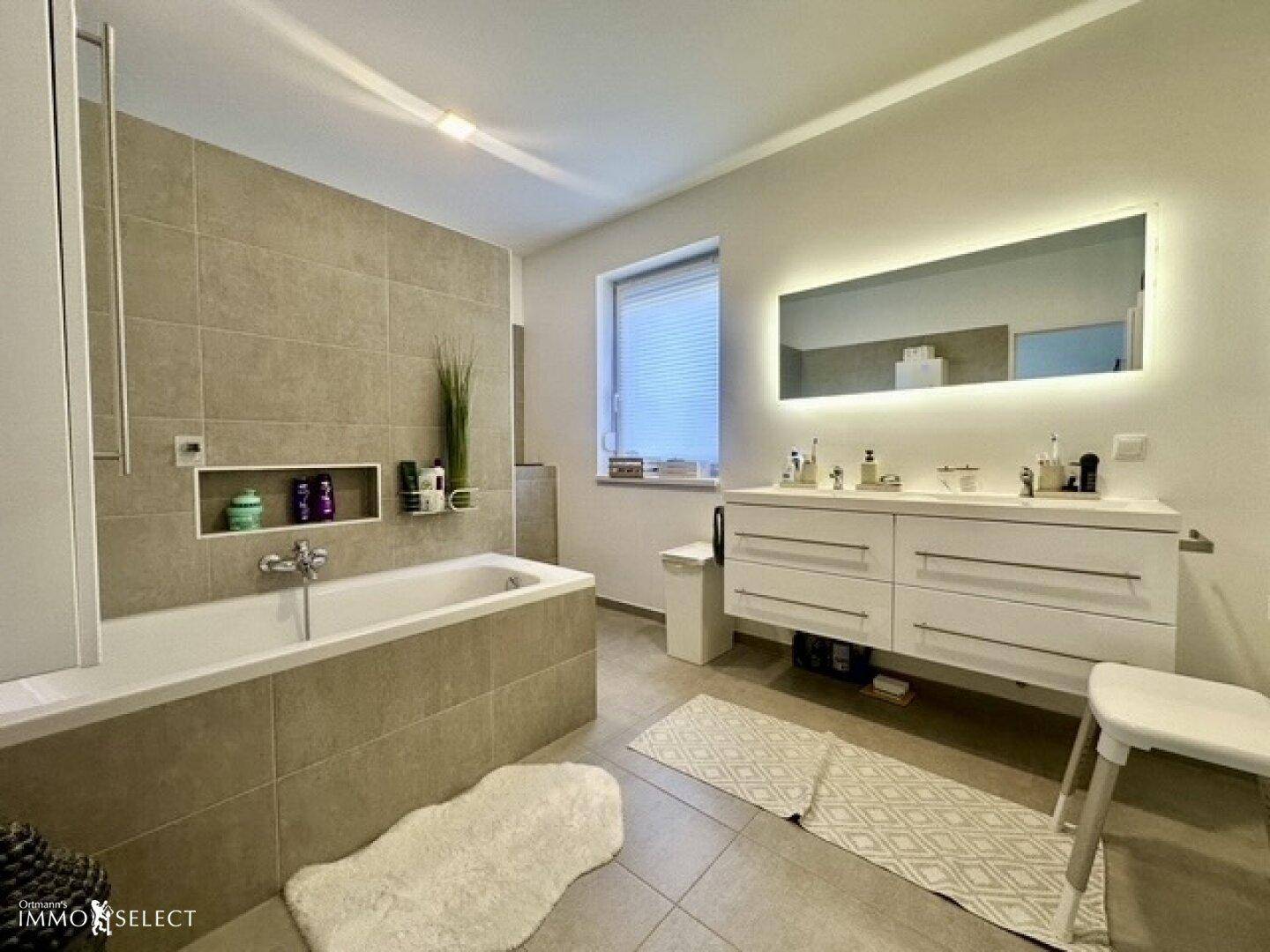 Modernes Badezimmer mit Badewanne, Doppelwaschbecken und großem beleuchteten Spiegel, in hellen Grautönen gehalten.