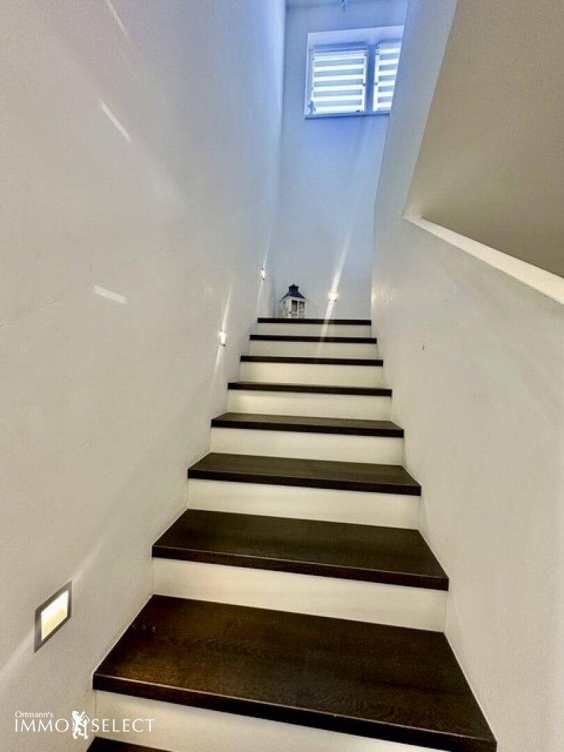 Elegante Treppe mit dunklen Holzstufen und weißem Geländer, beleuchtet durch Wandleuchten.