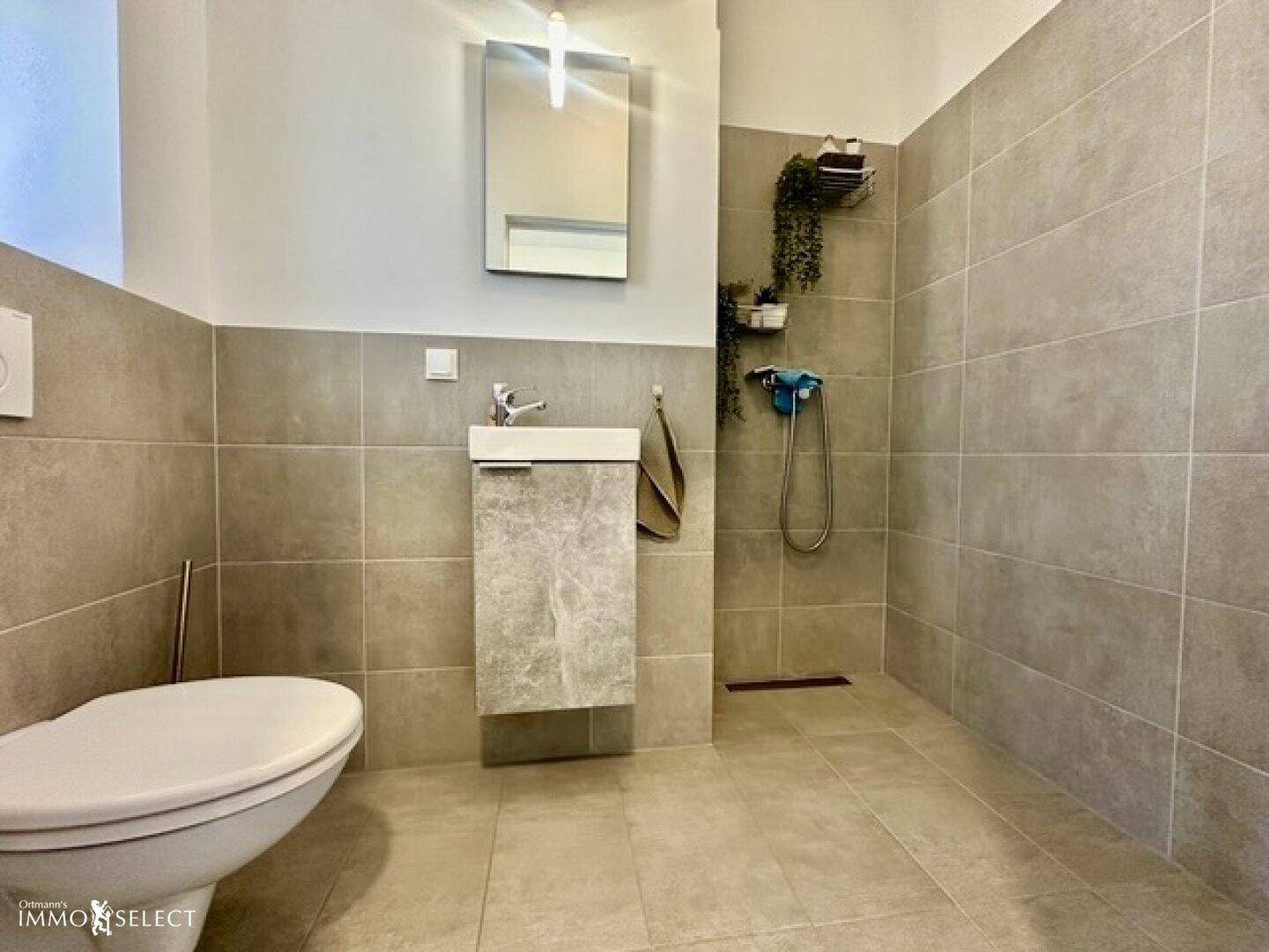 Modernes Badezimmer mit grauen Fliesen, Dusche und wandhängendem WC.