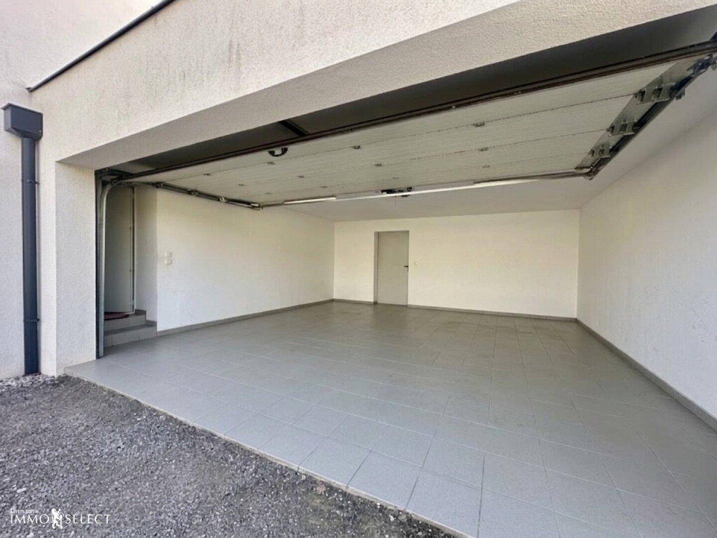 Geräumige, leere Garage mit gefliestem Boden und offenem Garagentor.