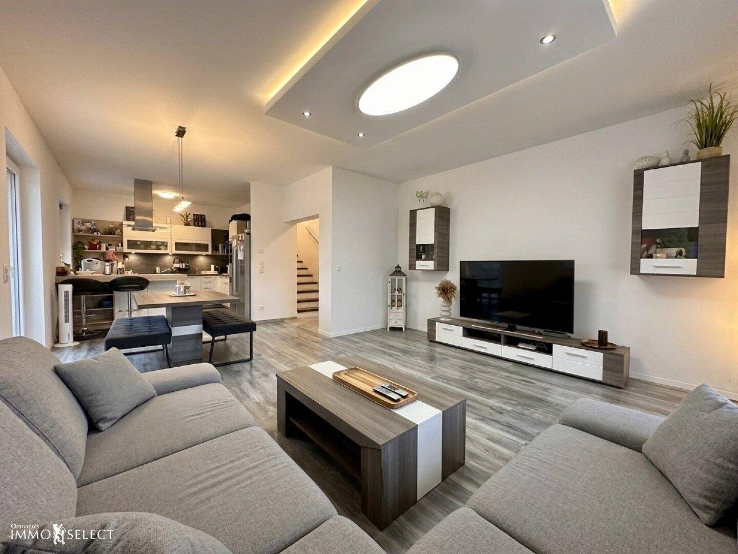 Offener Wohnbereich mit großem Sofa, Fernseher und moderner Deckenbeleuchtung, verbunden mit der Küche.