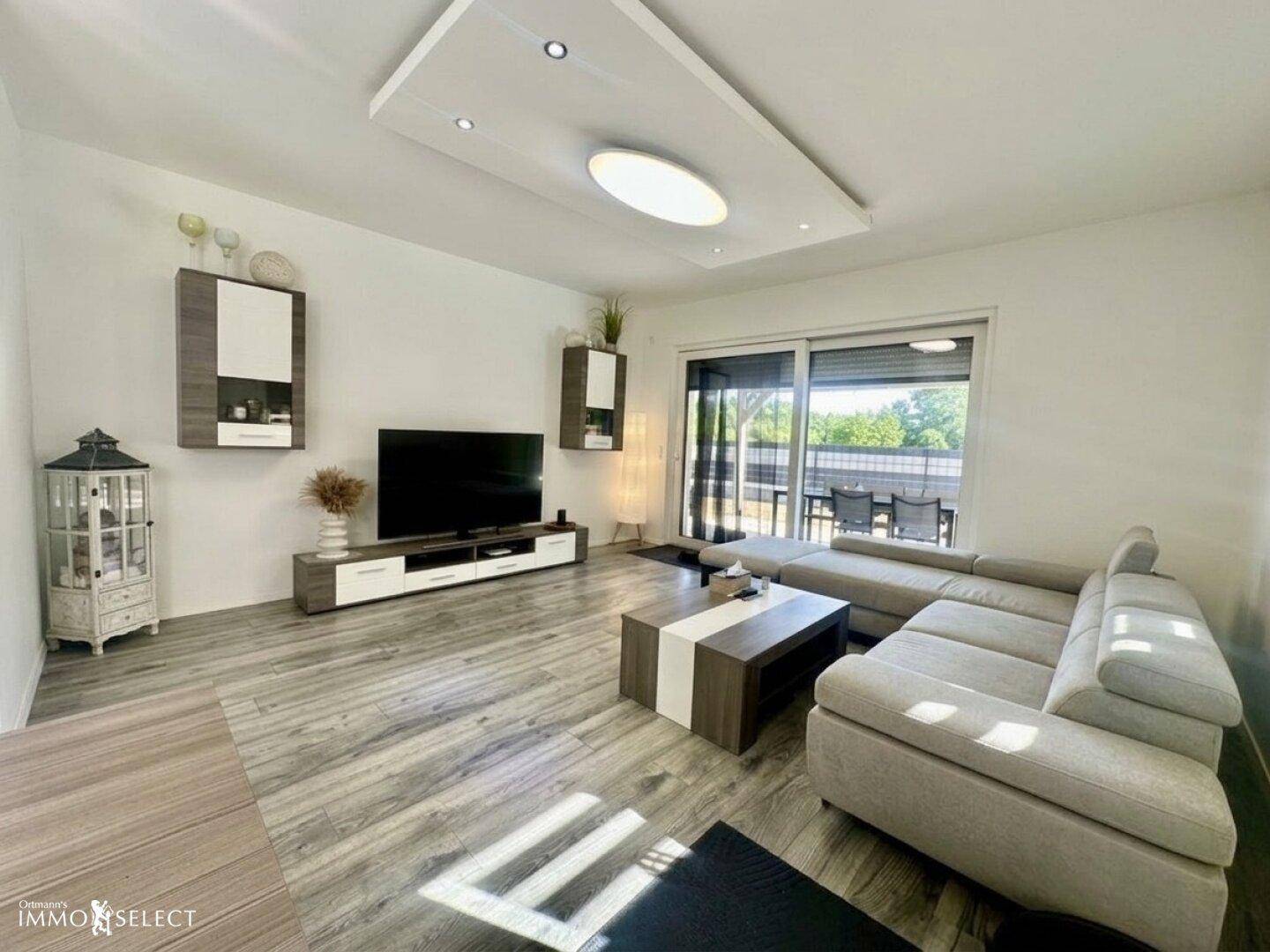 Heller Wohnbereich mit großem Sofa, Fernseher und Zugang zu einer Terrasse durch eine Glasschiebetür.