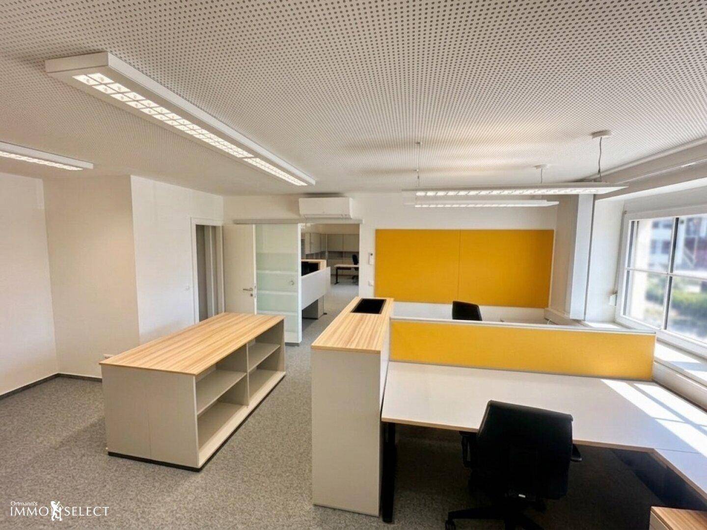 Helles Büro mit modernen Arbeitsplätzen, gelben Akzenten und großen Fenstern.