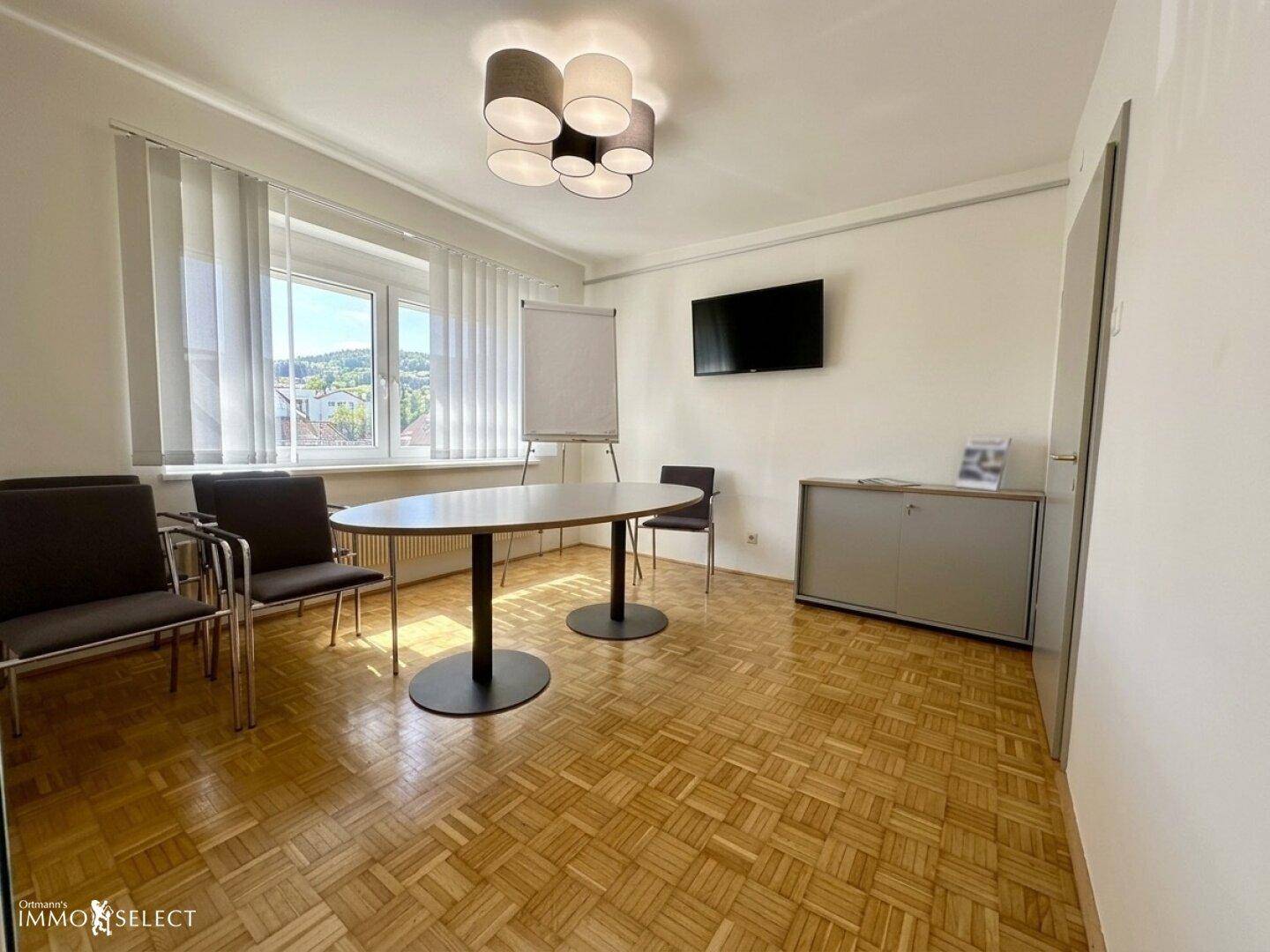 Besprechungsraum mit Parkettboden, ovalem Tisch, Stühlen und Wandfernseher.