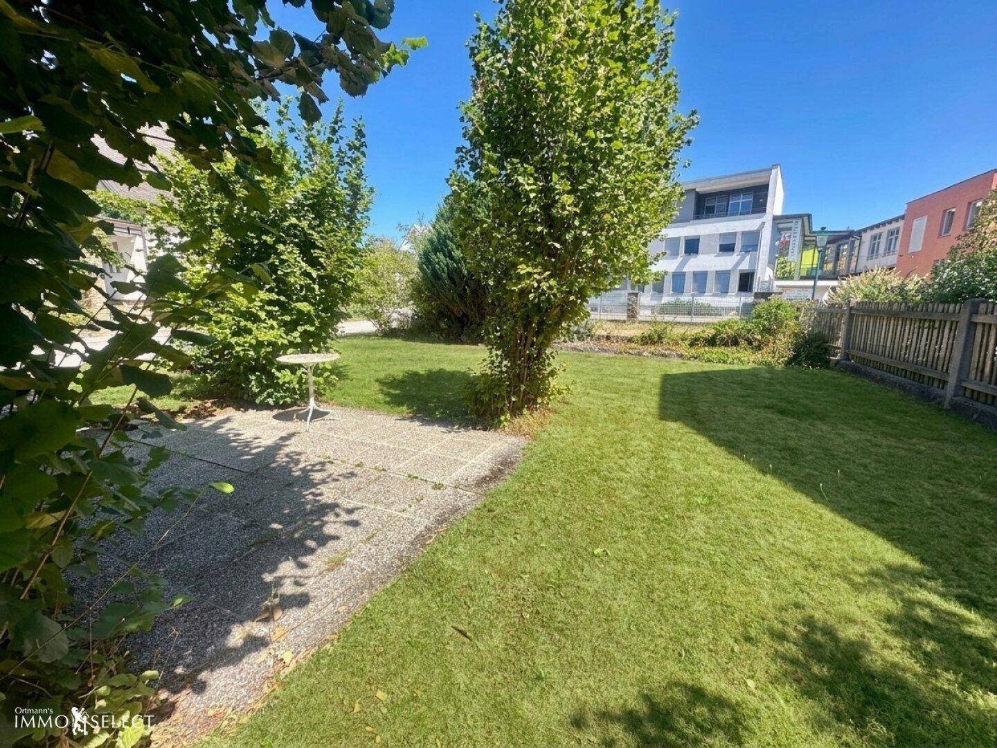 Sonniger Garten mit Rasenfläche, kleiner gepflasterter Terrasse und üppiger Bepflanzung.