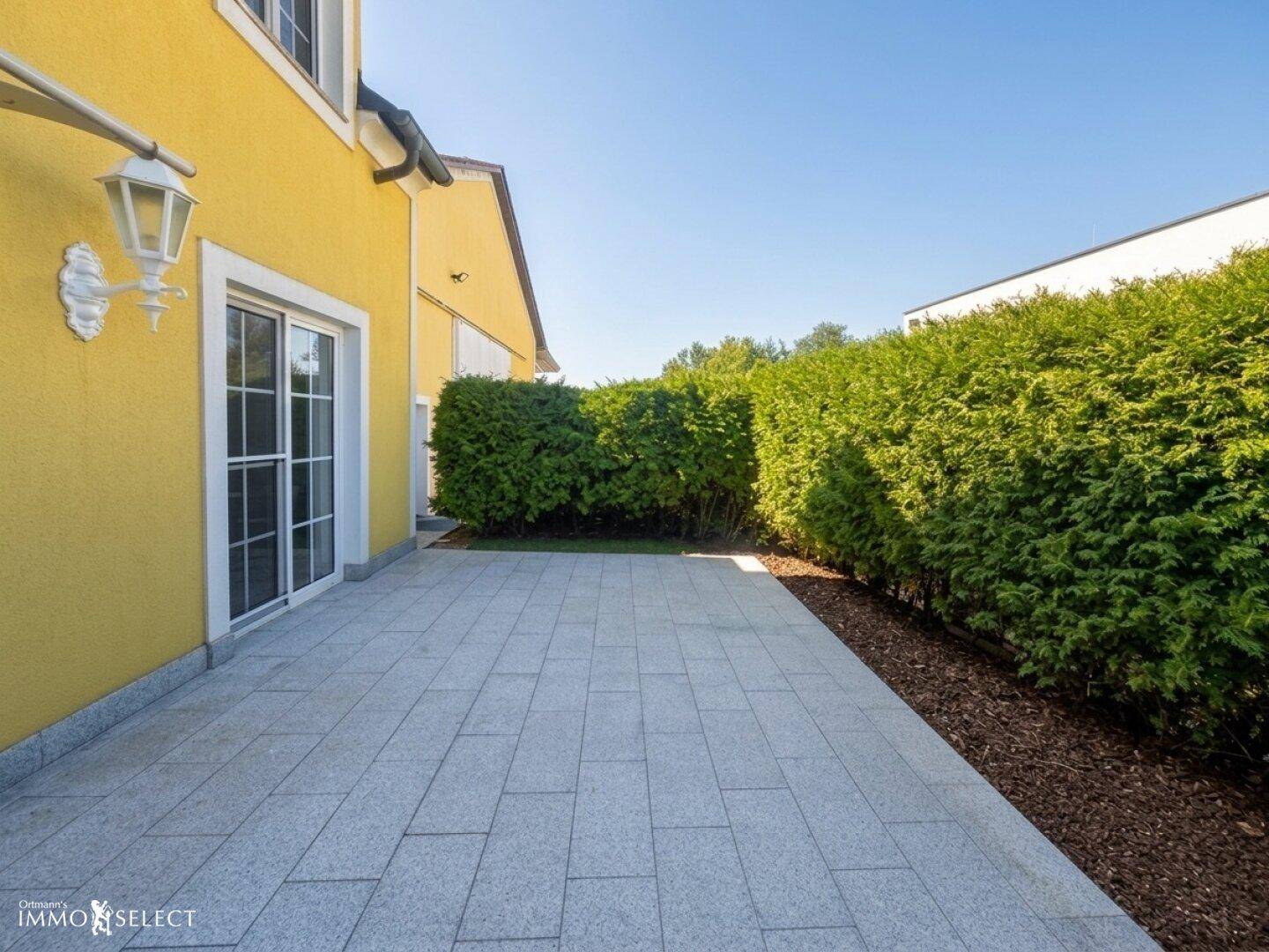 Gepflasterte Terrasse am Haus mit Hecke und Zugang zum Innenbereich.
