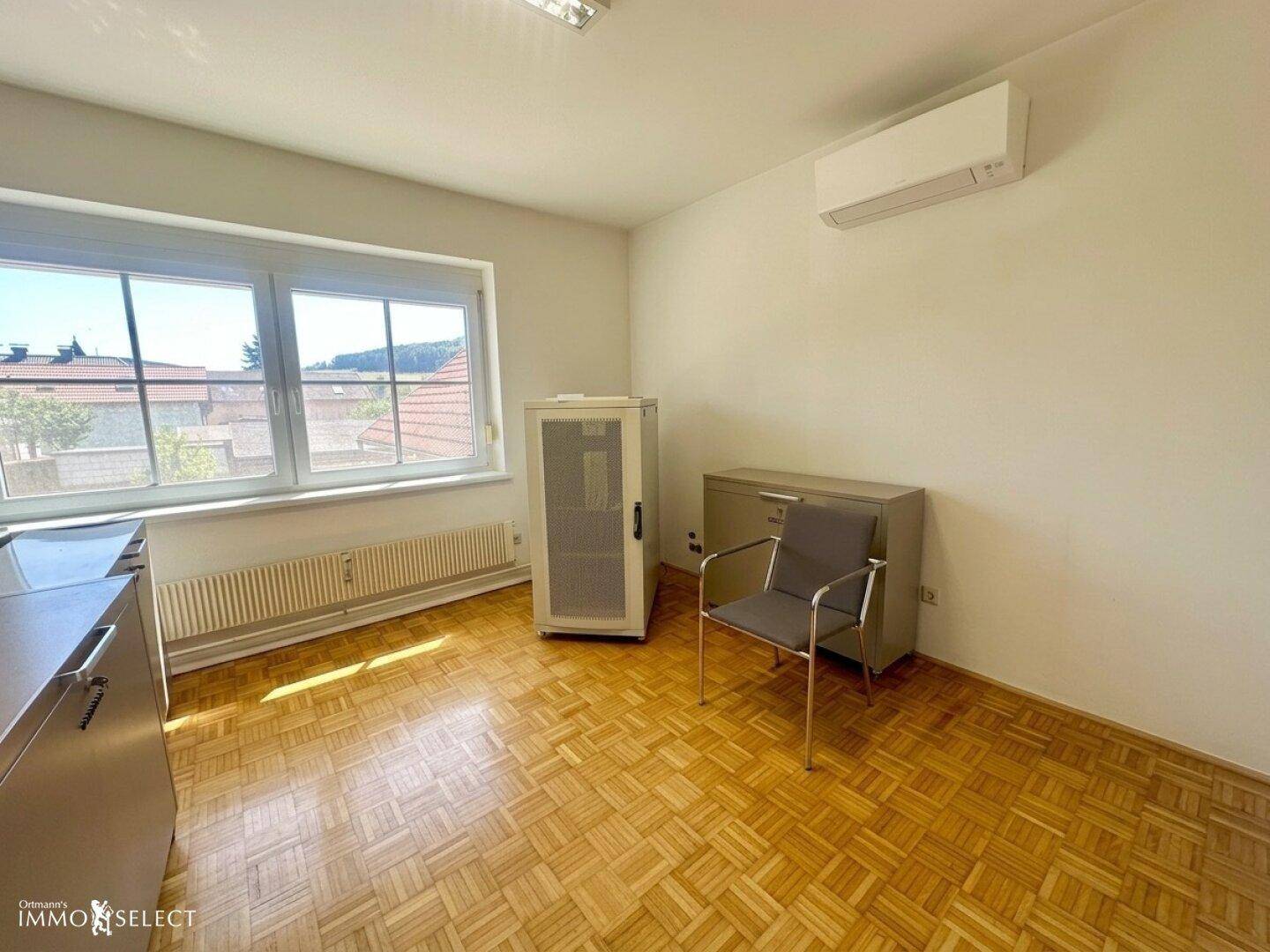 Helles Bürozimmer mit Parkettboden, Klimaanlage und Blick ins Grüne durch das Fenster.