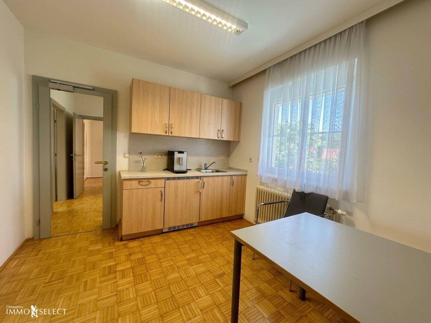Kleine Küchenzeile mit Holzschränken, Spüle und einem Fenster mit Vorhängen.