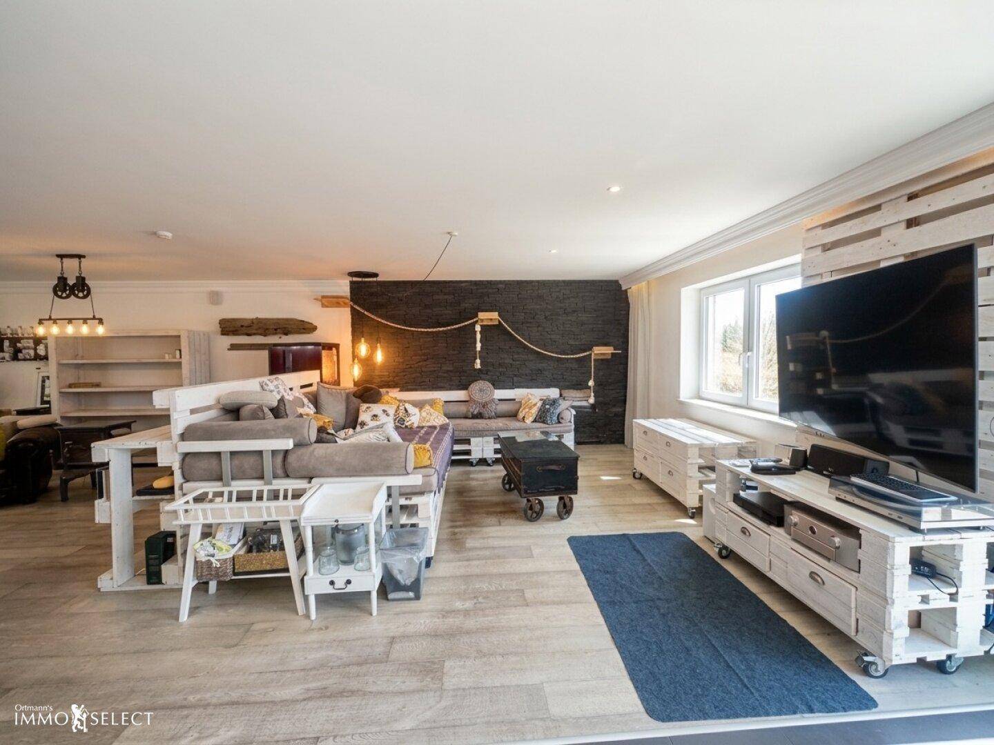 Gemütliches Wohnzimmer mit großem Sofa, Fernseher auf Palettenmöbeln und hellem Holzboden.