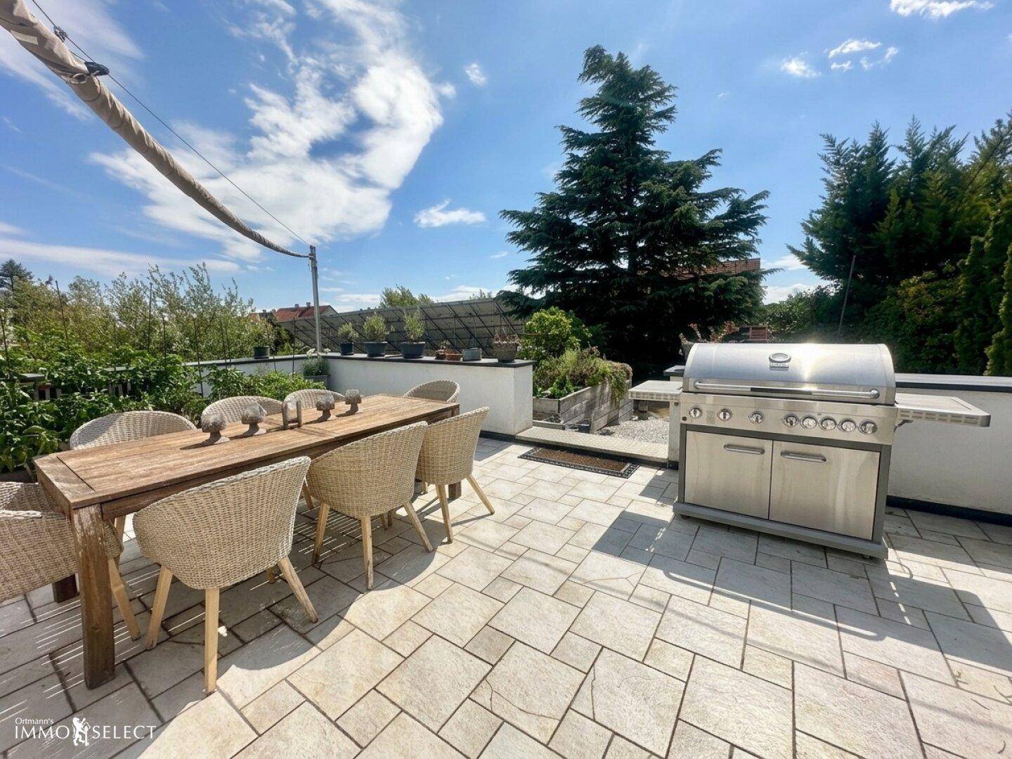 Große Außenterrasse mit Esstisch, Stühlen und einem modernen Grillbereich unter blauem Himmel.