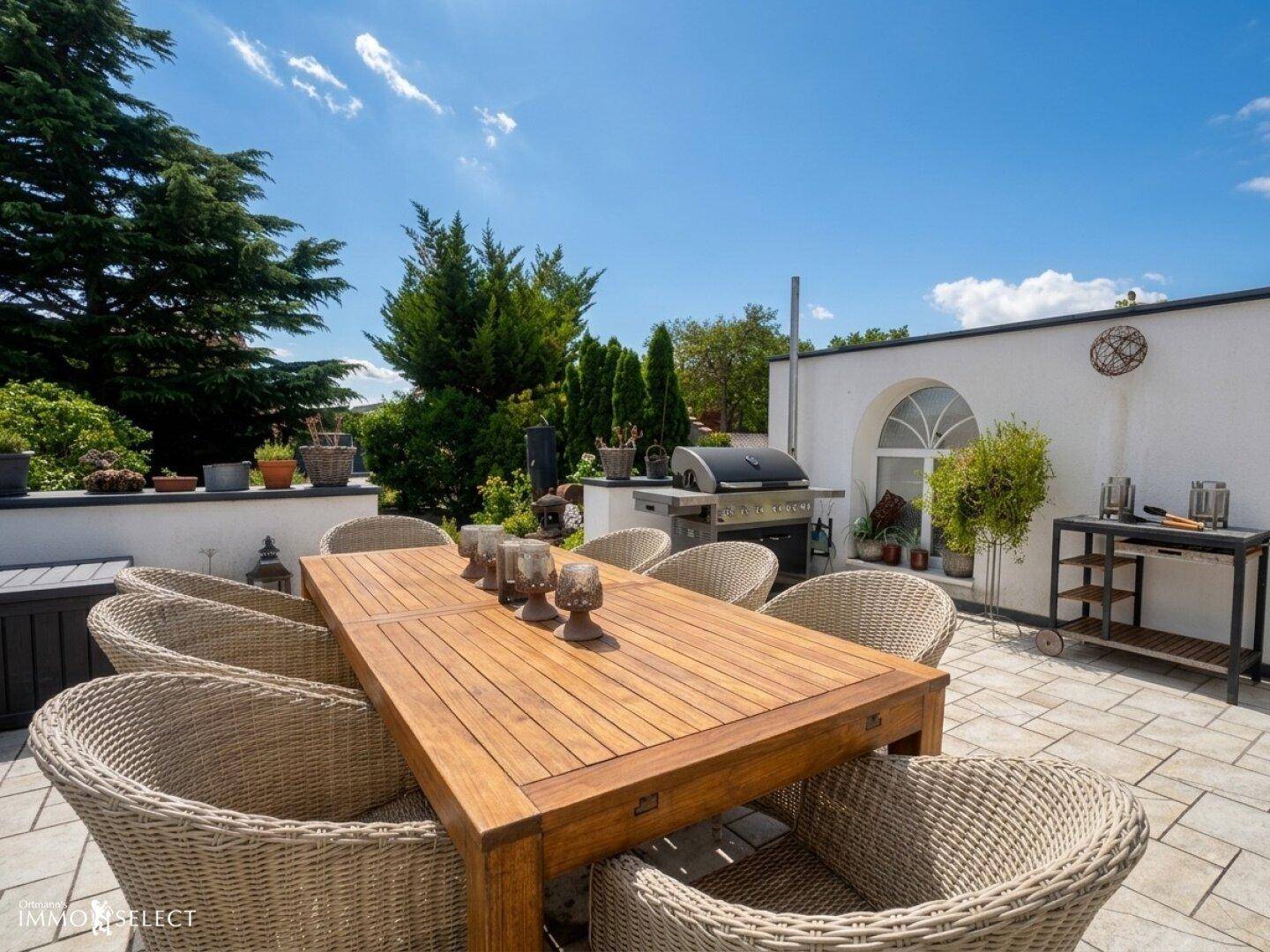 Detailansicht der sonnigen Terrasse mit Holztisch, Korbstühlen und Grillstation.