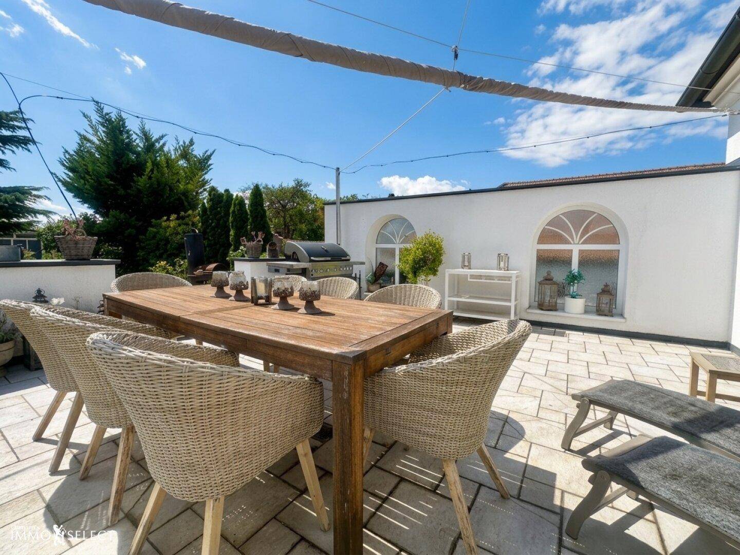 Große gepflasterte Terrasse mit Esstisch, Korbstühlen und Grillbereich unter blauem Himmel.