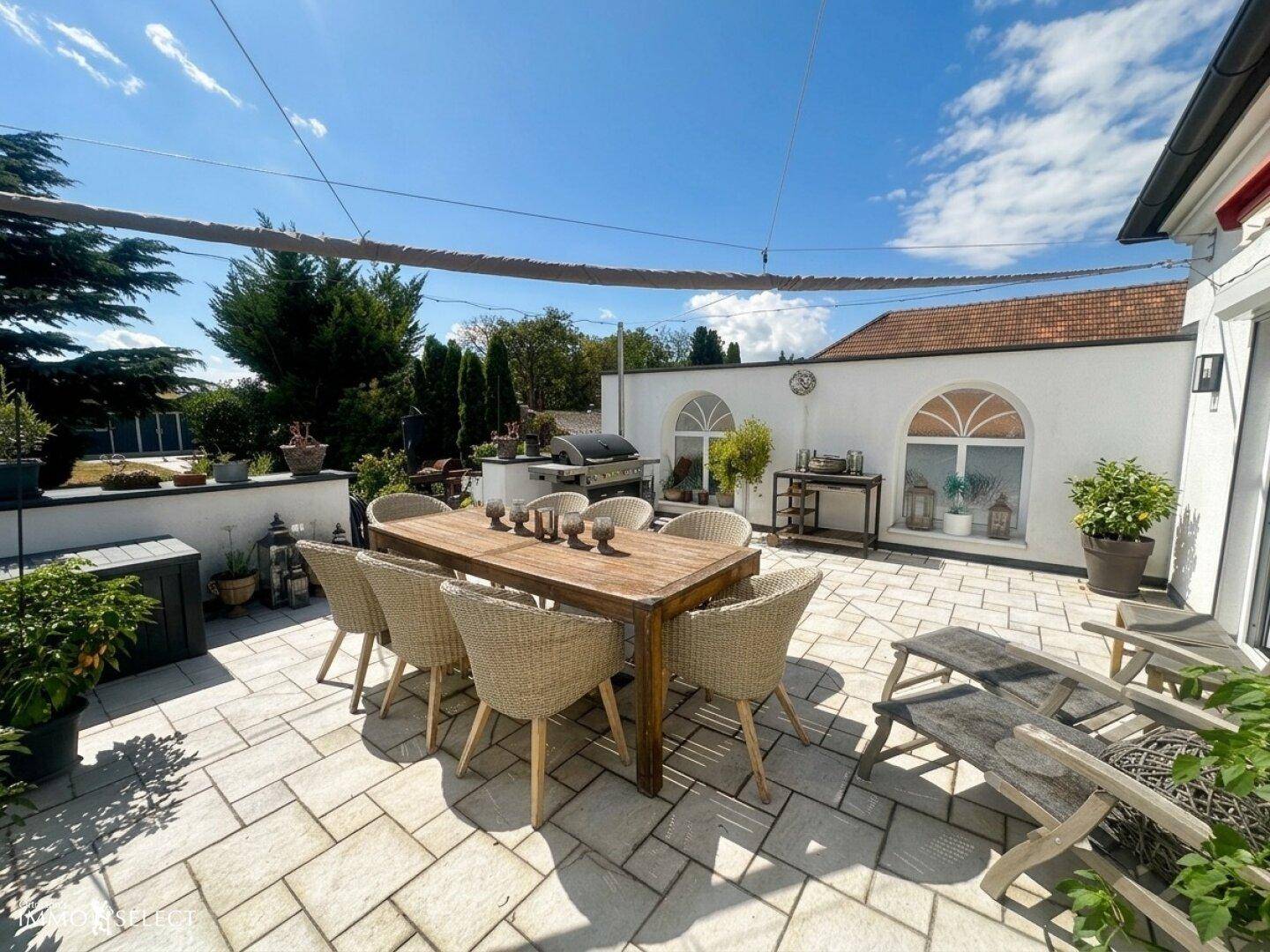 Sonnige Terrasse mit großem Holztisch, Korbstühlen und Grillplatz im Freien.