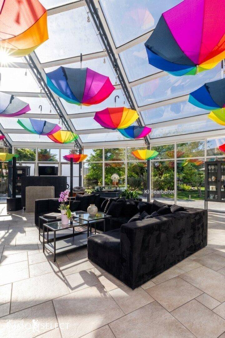 Lichtdurchfluteter Wintergarten mit bunten Regenschirmen, bequemen Sofas und Blick in den Garten.