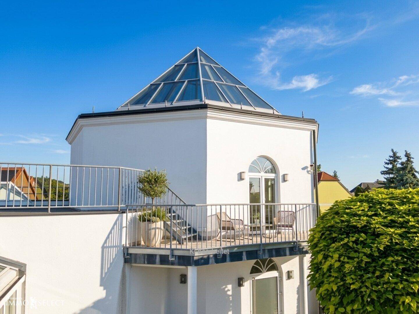 Weißes Gebäude mit pyramidenförmigem Glasdach und einem kleinen Balkon mit Sitzgelegenheiten.