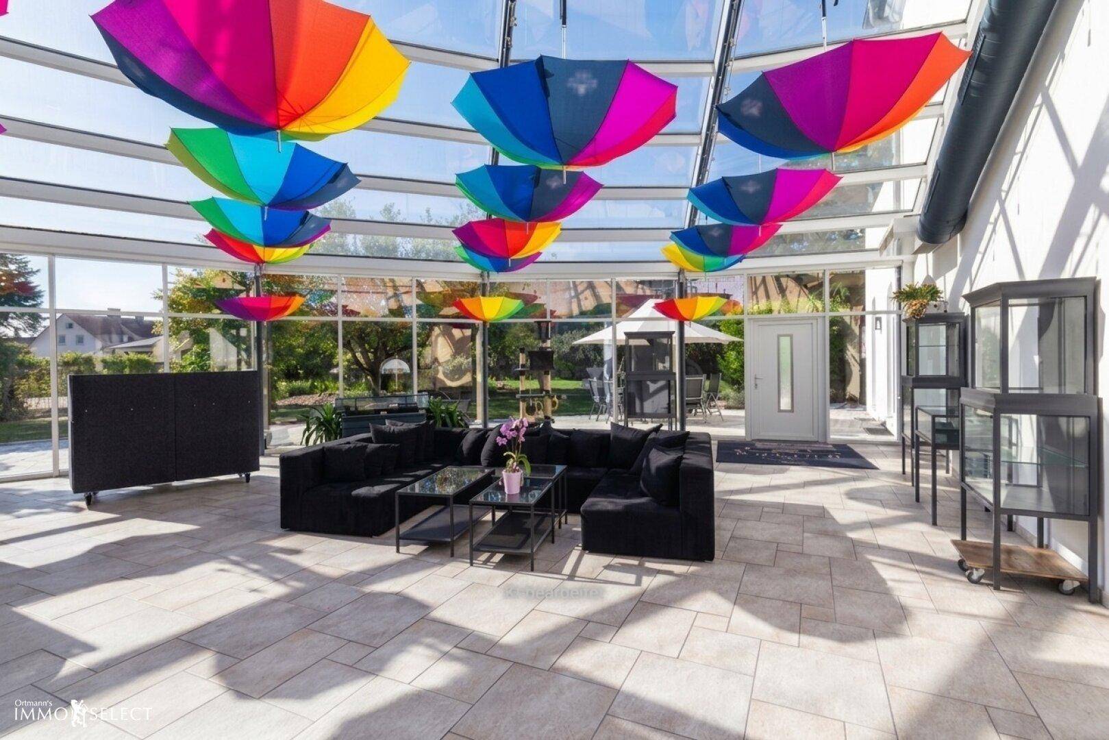 Lichtdurchfluteter Wintergarten mit bunten Regenschirmen, bequemen Sofas und Blick in den Garten.