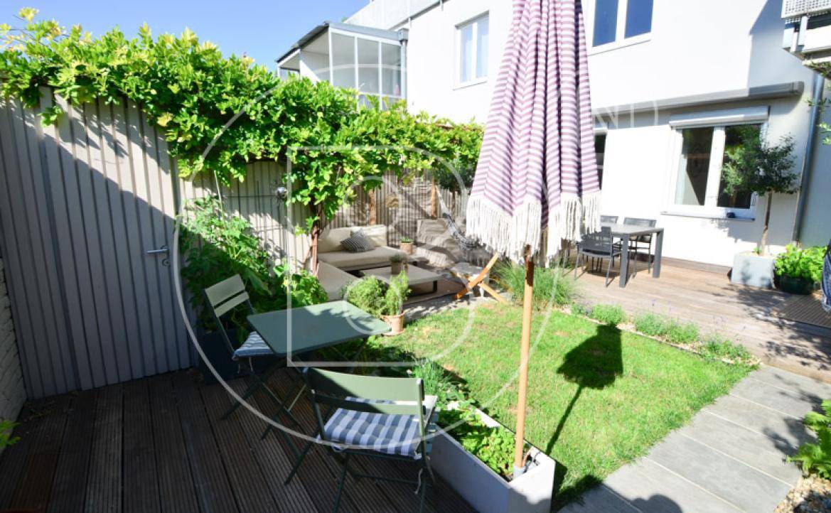Sonnige Holzterrasse mit Gartenmöbeln, Sonnenschirm und kleiner Rasenfläche.