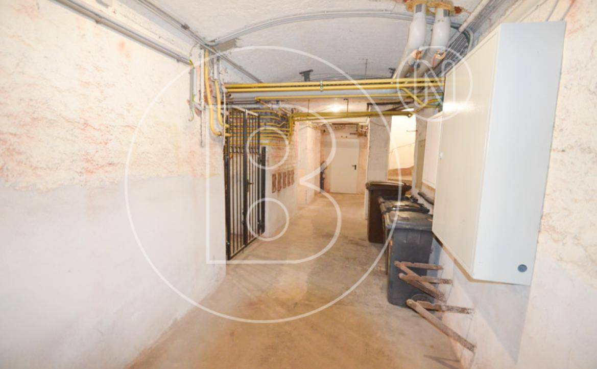 Kellerflur mit gelben Rohrleitungen an der Decke, verschließbaren Abteilen und einem Technikschrank.