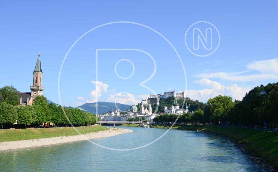 Malerischer Blick auf die Stadt Salzburg mit Fluss, Bäumen und der Festung Hohensalzburg im Hintergrund.