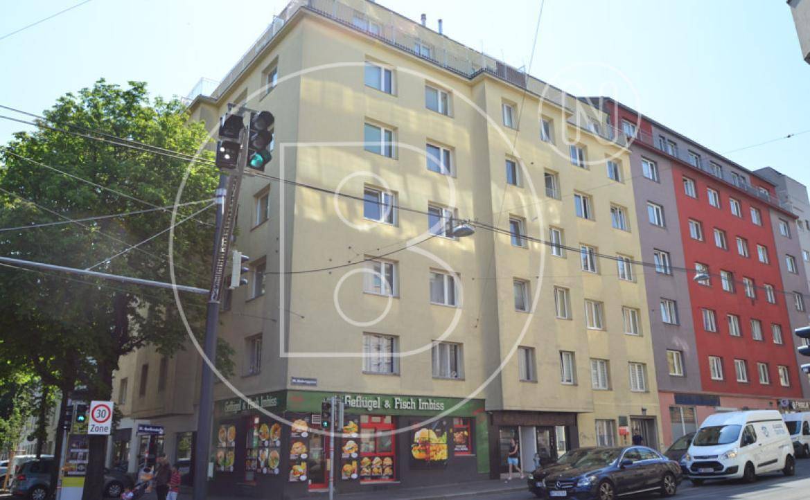 Eckgebäude mit gelber Fassade und Gewerbeflächen im Erdgeschoss, umgeben von städtischer Bebauung.