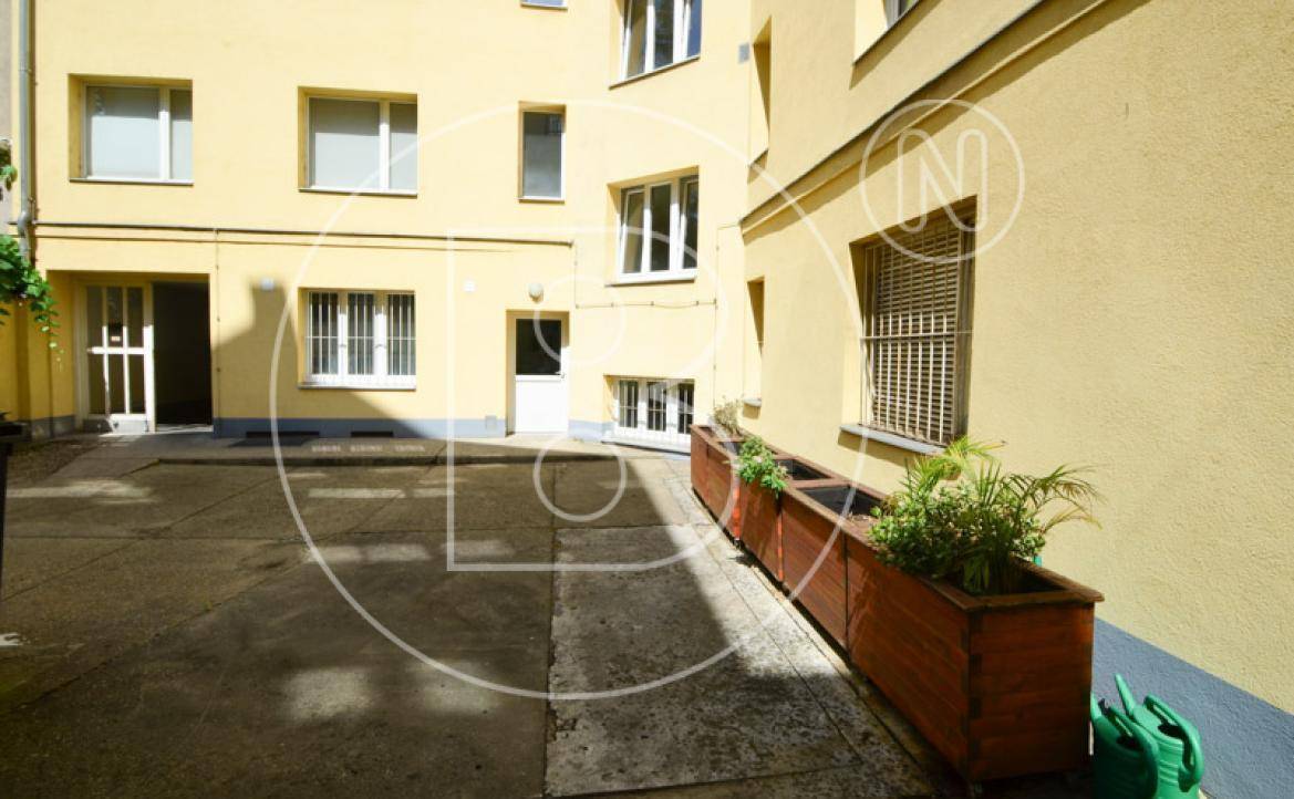 Ruhiger Innenhof mit gelber Fassade, bepflanzten Holzkübeln und einer Gießkanne.