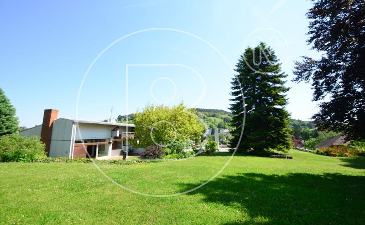 Ansicht des Hauses von der weitläufigen Rasenfläche mit Bäumen und Hügeln im Hintergrund.
