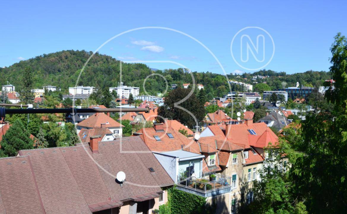 Panoramablick über die Dächer einer Wohngegend mit grünen Hügeln im Hintergrund unter blauem Himmel.