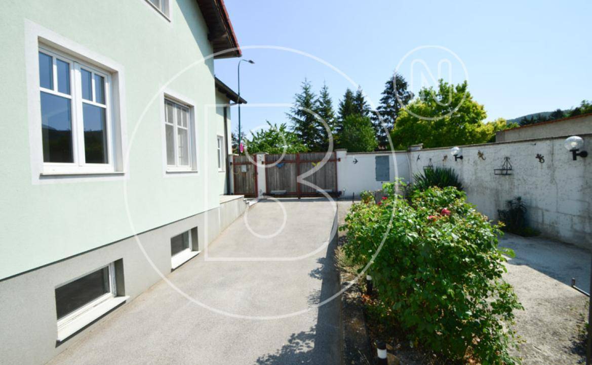 Seitenansicht des Hauses mit Rampe, Gartenbereich und alter Mauer unter blauem Himmel.