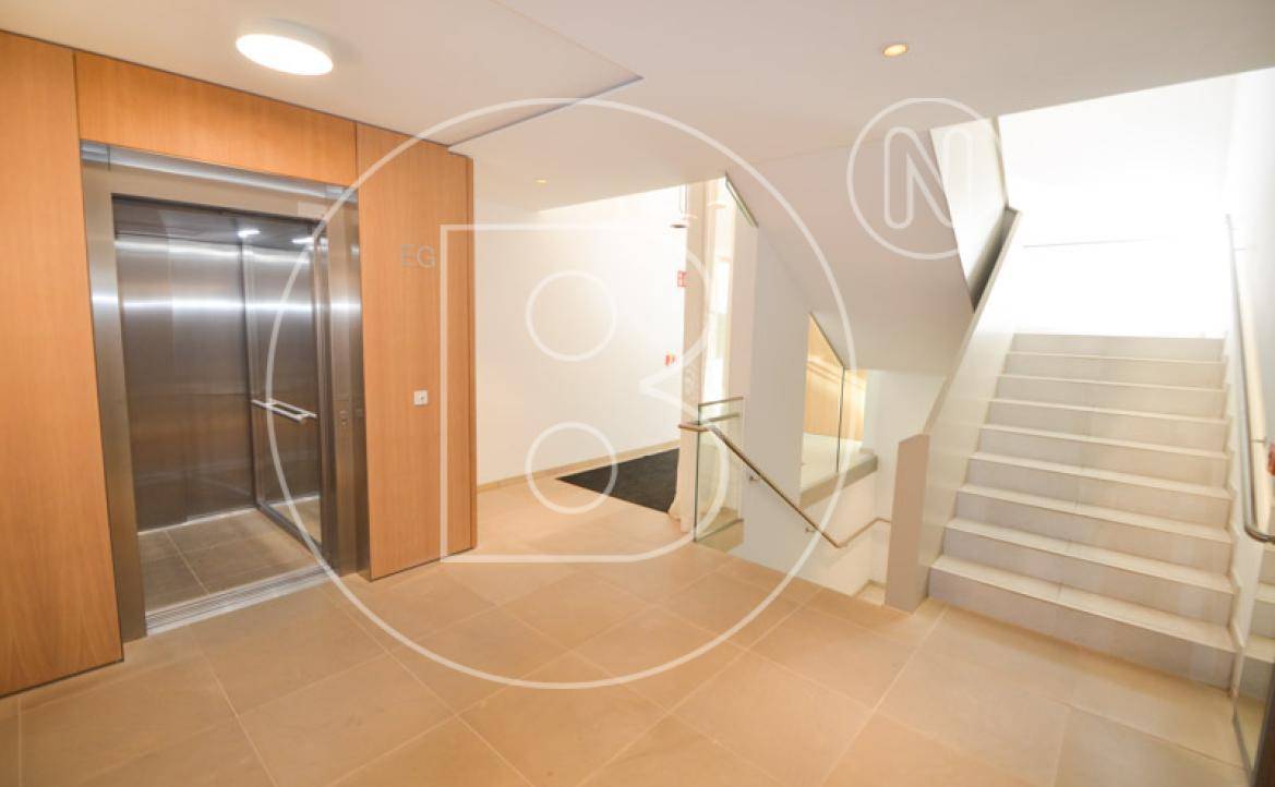 Heller Eingangsbereich mit modernem Aufzug und einer eleganten Treppe mit Glasgeländer.