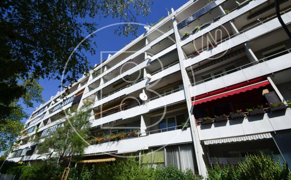 Fassade eines Apartmentgebäudes mit vielen Balkonen, Markisen und üppiger Bepflanzung unter klarem Himmel.