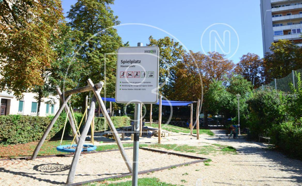 Öffentlicher Spielplatz mit Schaukeln und Sandbereich, umgeben von Bäumen und einem Hinweisschild.