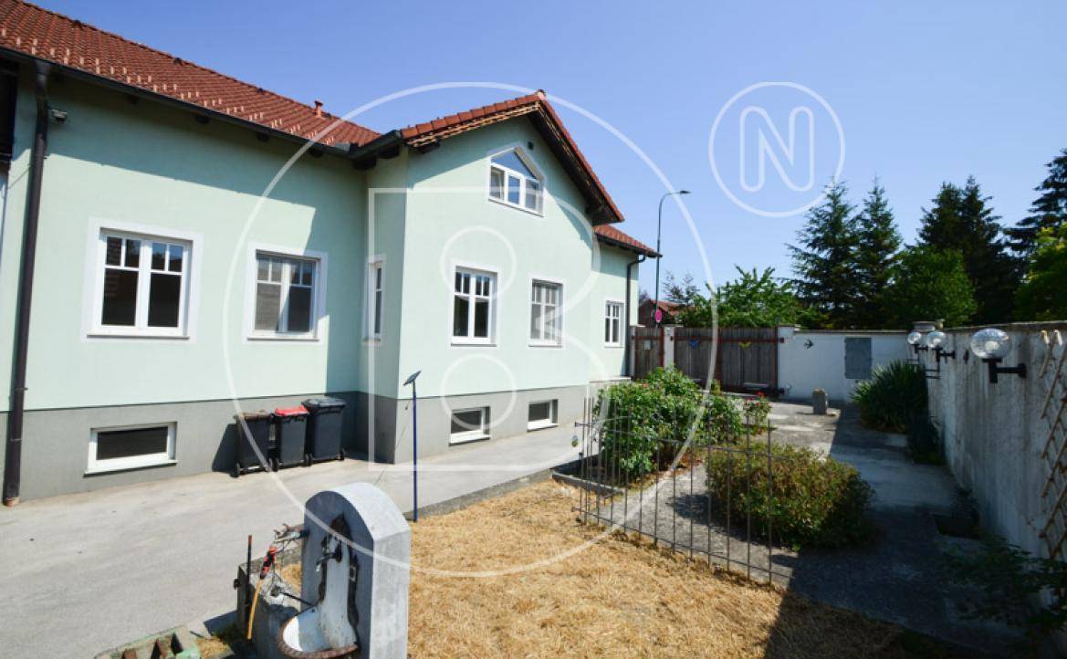 Seitenansicht des Hauses mit Gartenbereich, alter Mauer und Bäumen unter blauem Himmel.