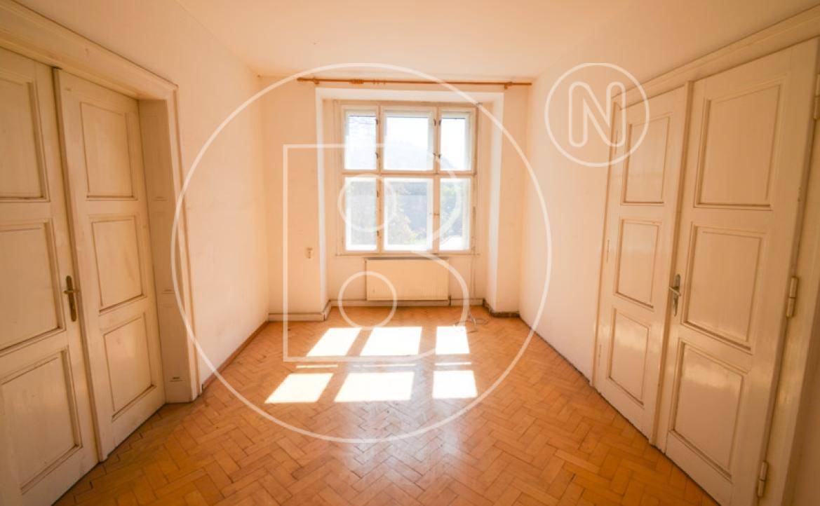 Ein leerer Raum mit Fischgrätparkett, zwei hohen Türen und einem Fenster, das viel Licht hereinlässt.