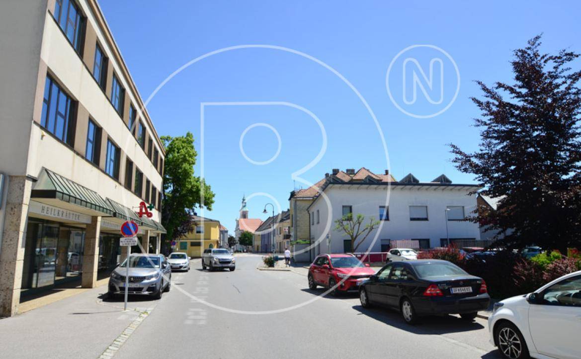 Belebtes Stadtbild mit Gebäuden, parkenden Autos und einer Straße unter klarem blauem Himmel.