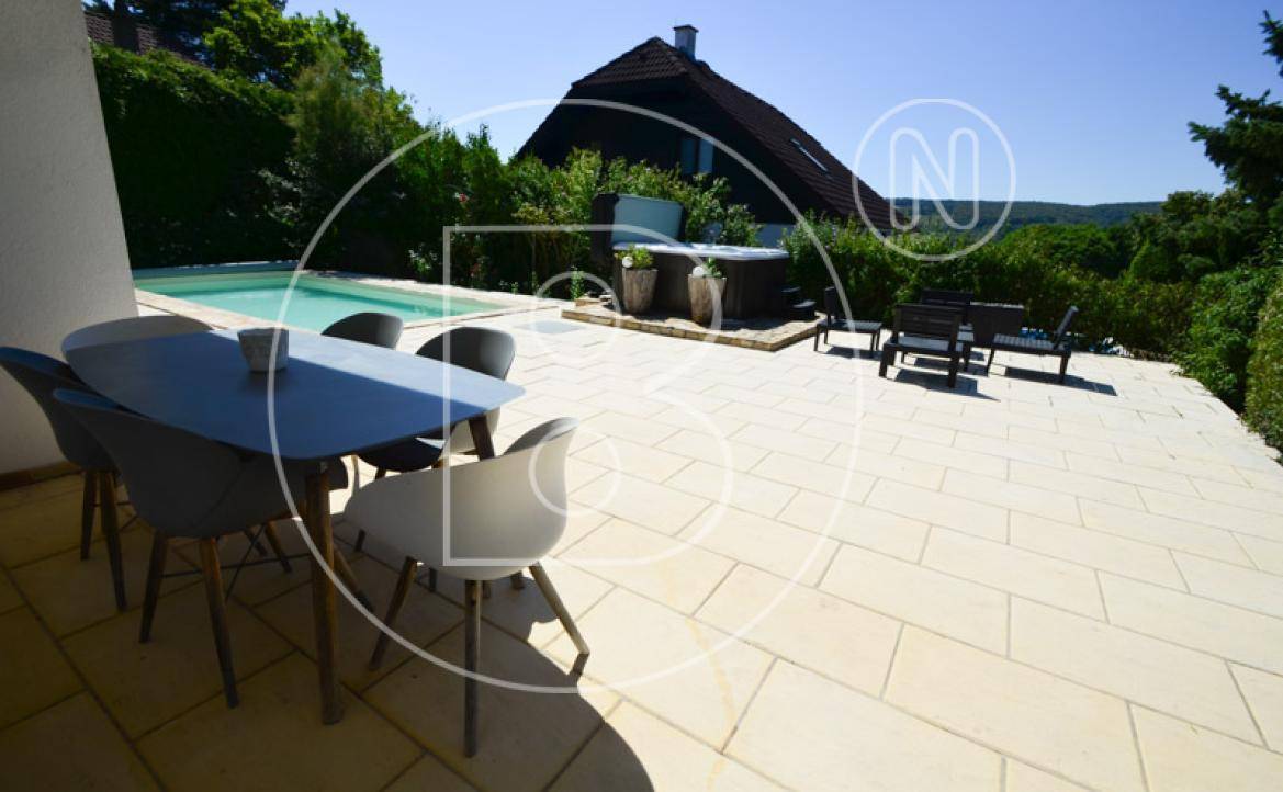 Große, helle Terrasse mit Esstisch und Stühlen, umgeben von Grün und mit Pool im Hintergrund.