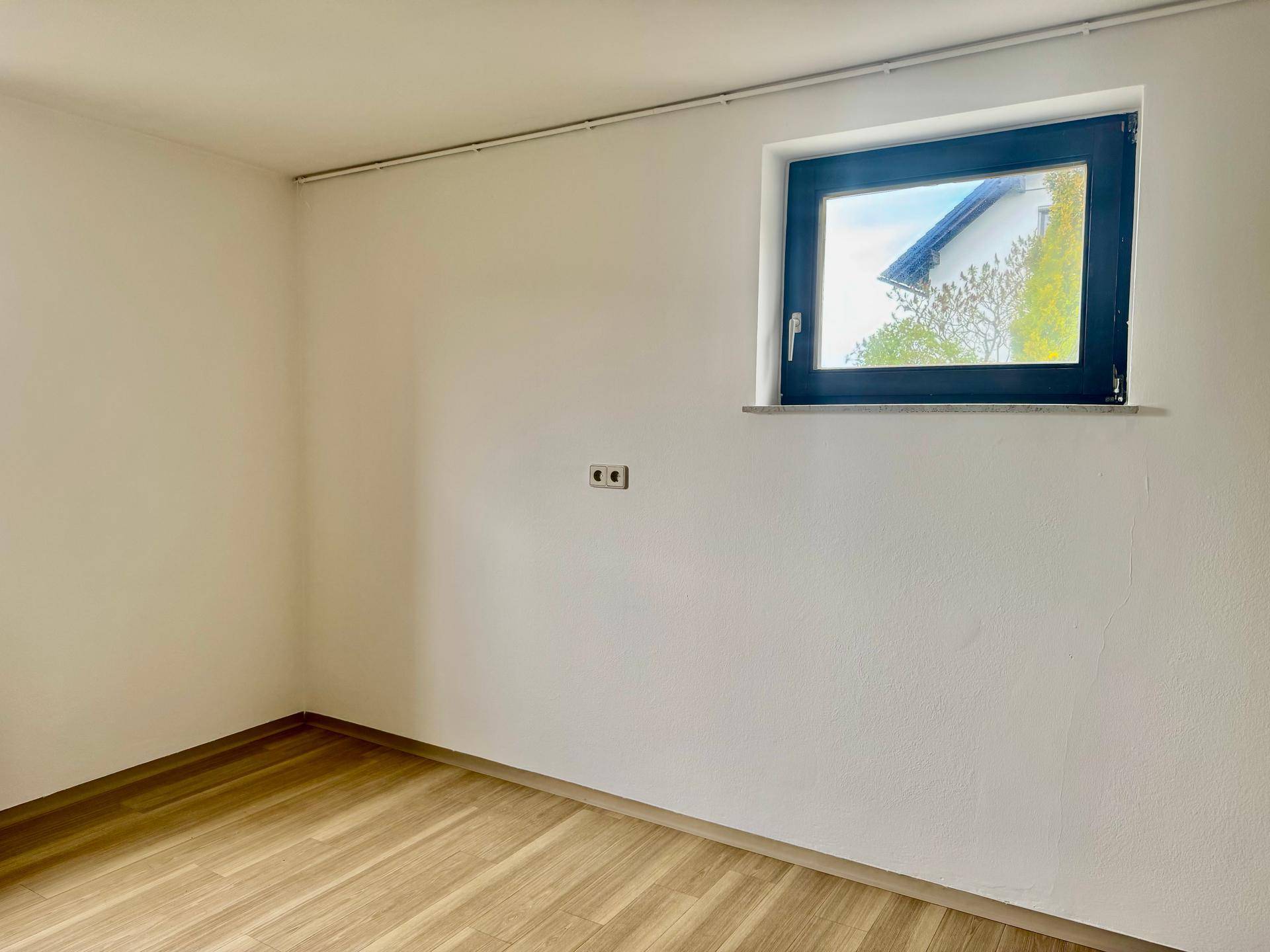 Kleines, helles Zimmer mit weiß gestrichenen Wänden, Holzfußboden und einem Fenster.