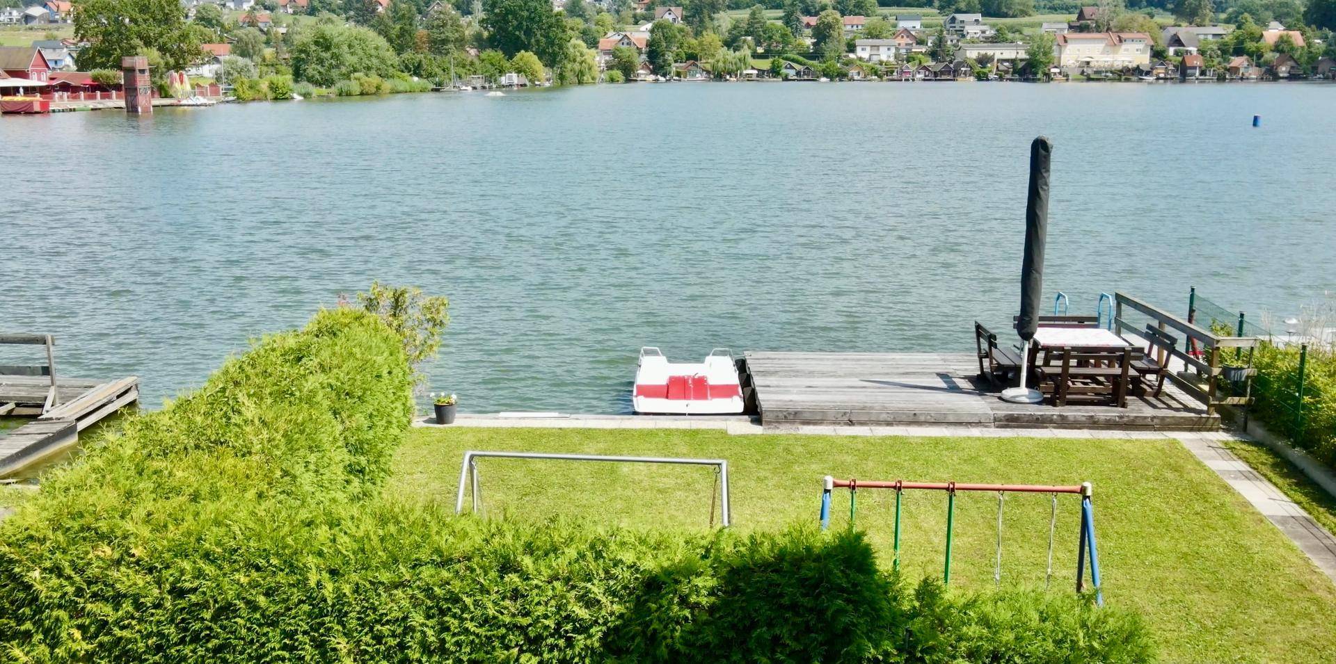 Weitläufiger Blick auf den See mit Steg, Schaukel und grüner Hecke im Vordergrund.