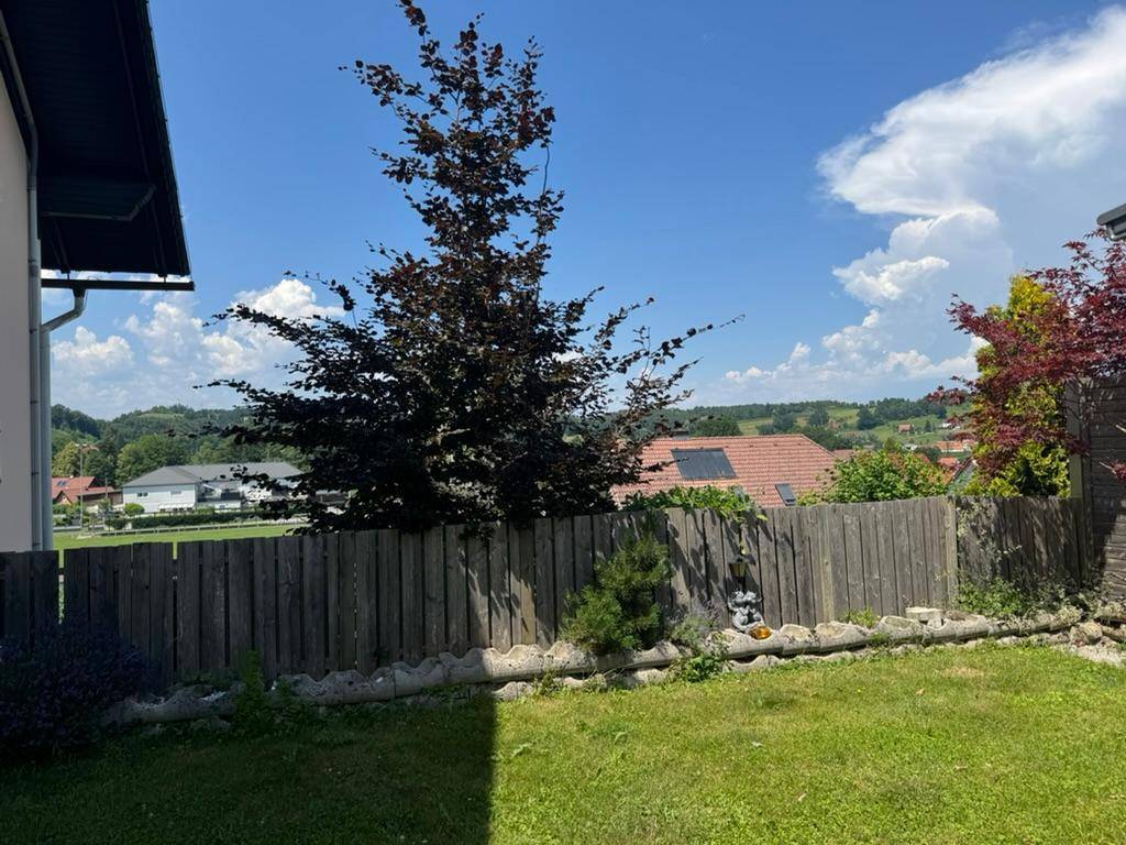 Blick aus dem Garten über einen Holzzaun auf die umliegende Landschaft mit Häusern und grünen Hügeln.