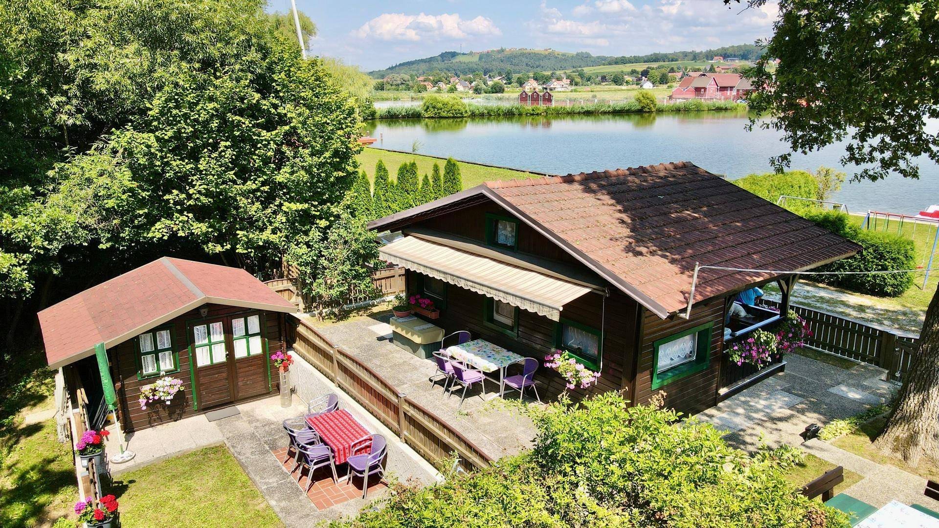 Vogelperspektive des Holzhauses mit Nebengebäude, Terrasse und See im Hintergrund.