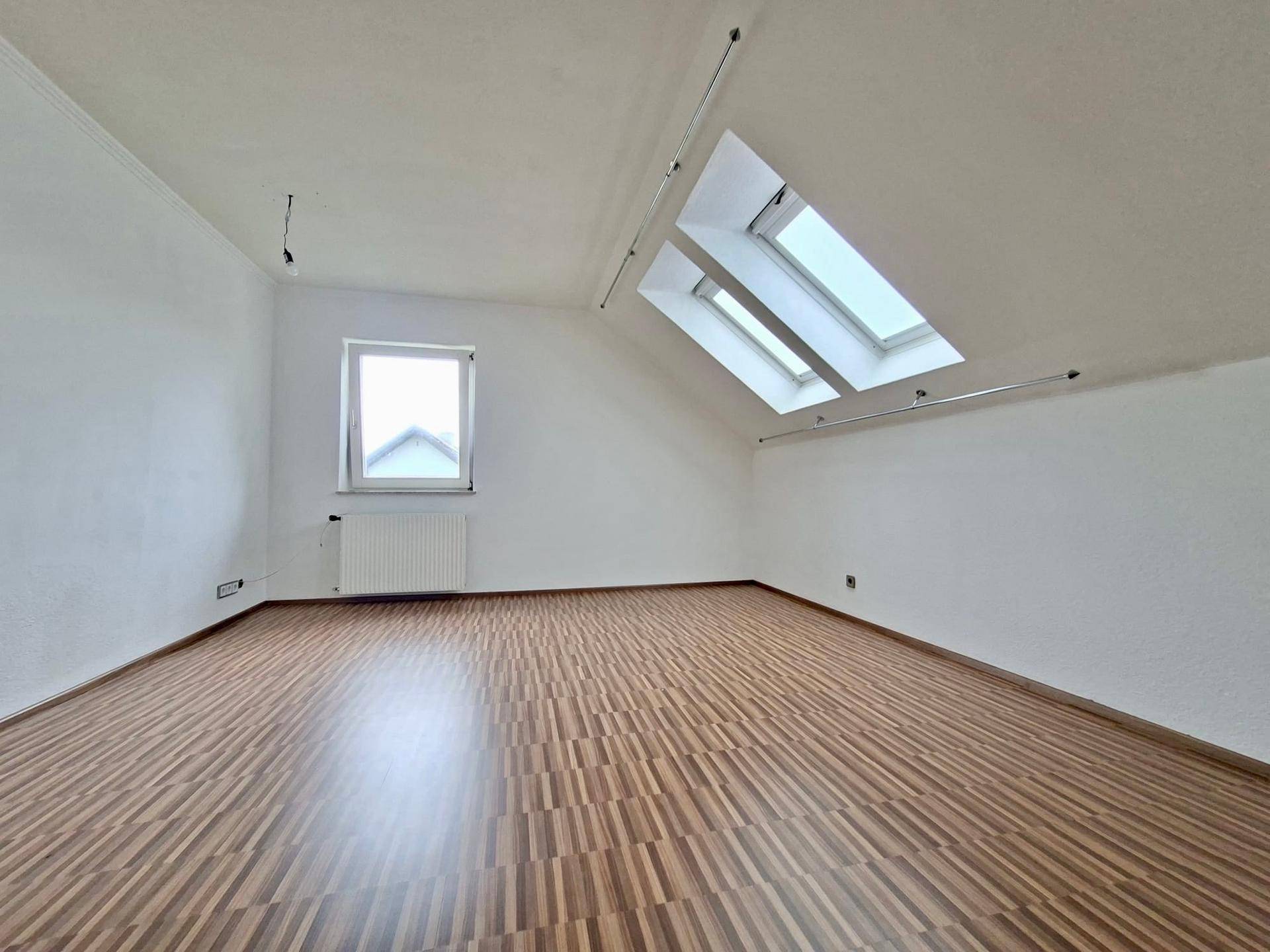 Dachzimmer mit schrägen Decken, Holzfußboden und zwei Dachfenstern für viel Tageslicht.