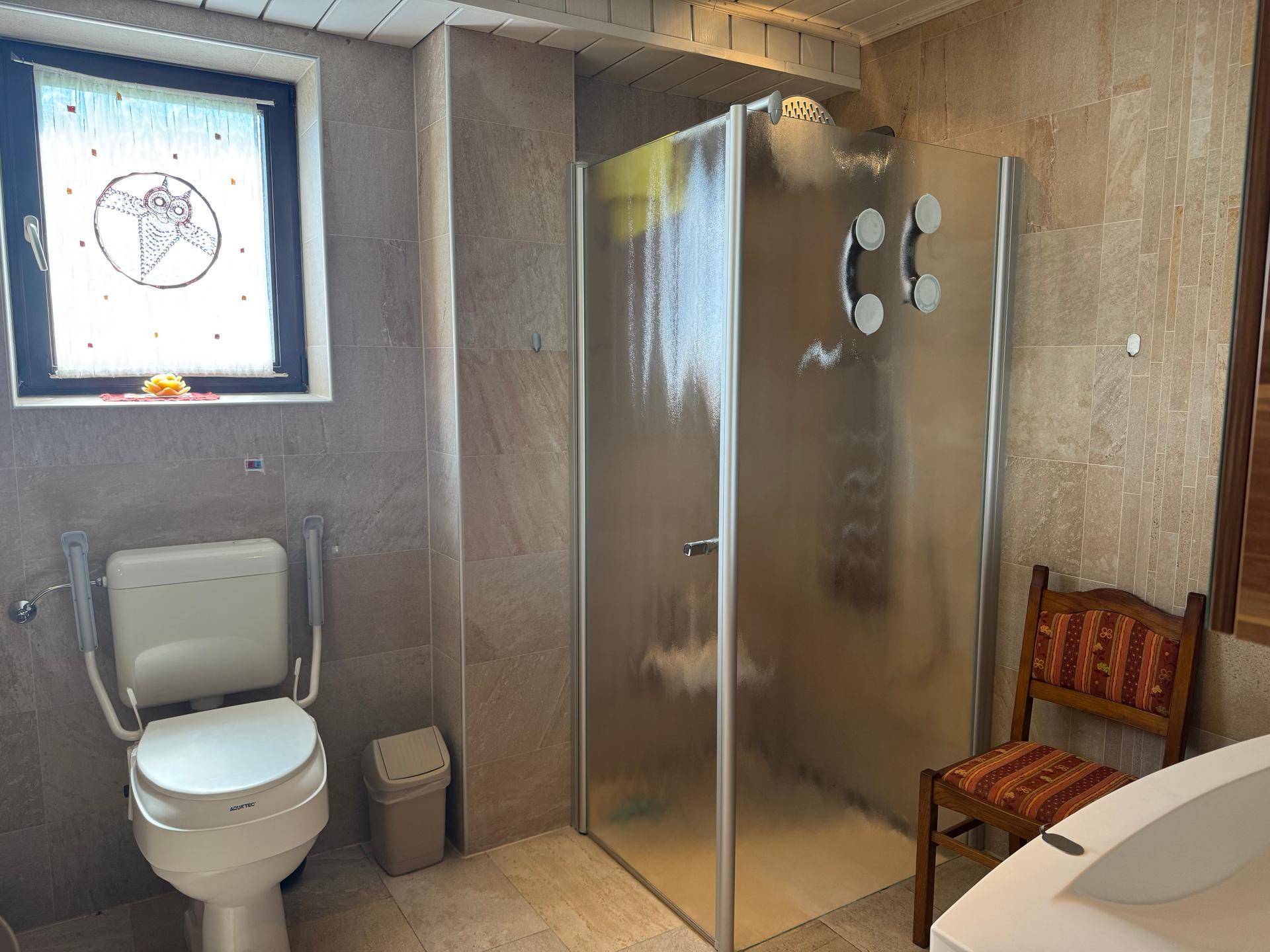 Badezimmer mit Duschkabine, WC und Fenster, ausgestattet mit hellen Fliesen.