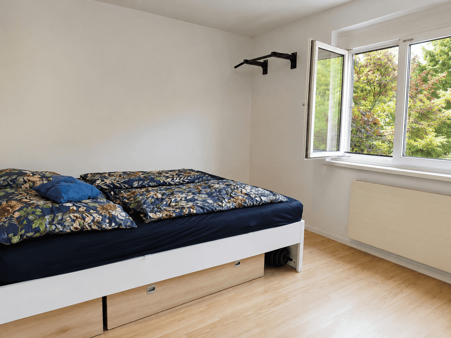 Helles Schlafzimmer mit Holzboden, großem Fenster und einem Bett mit Stauraum.