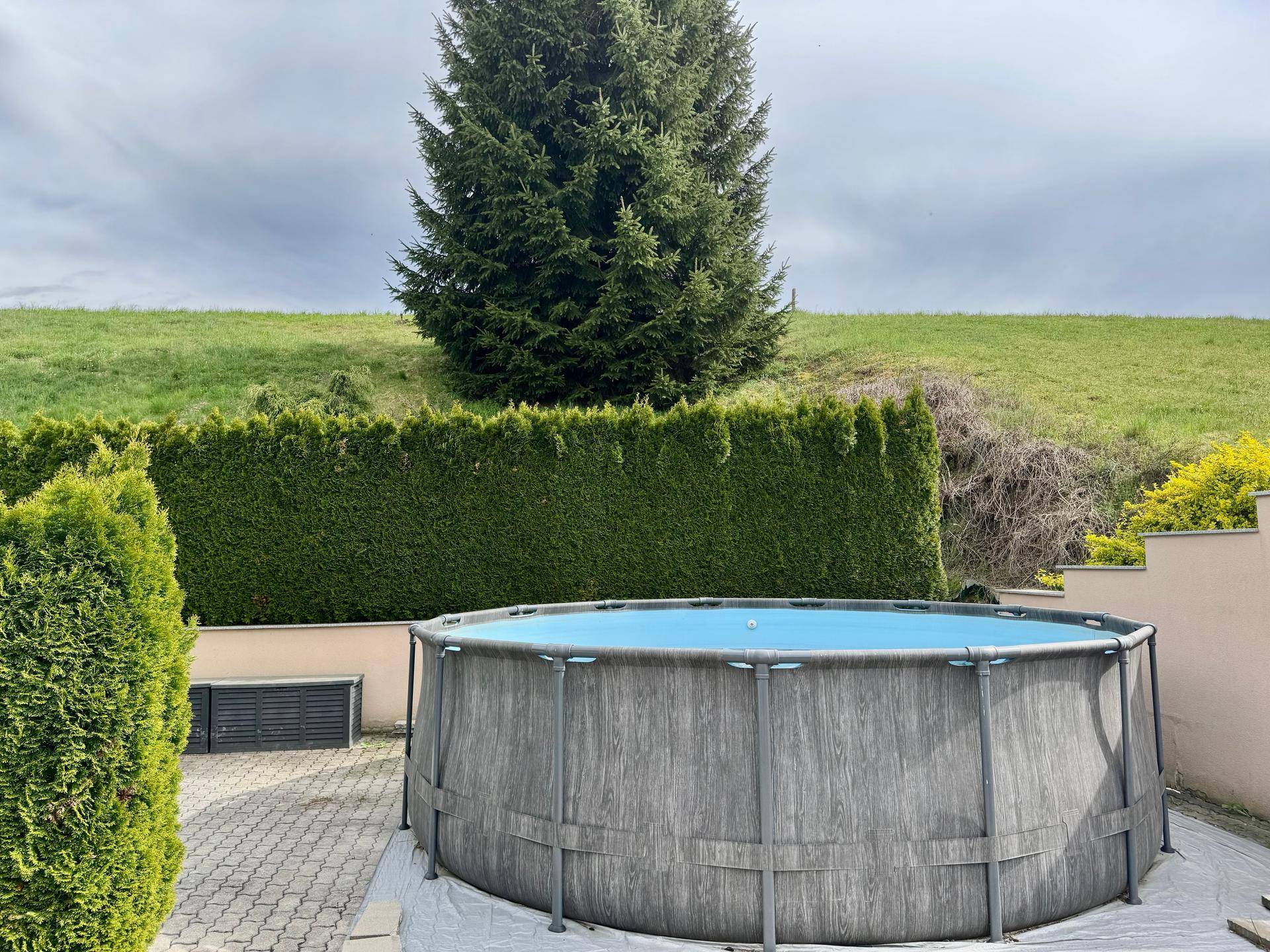 Großer Aufstellpool auf einer gepflasterten Fläche, umgeben von Hecken und Grünflächen.
