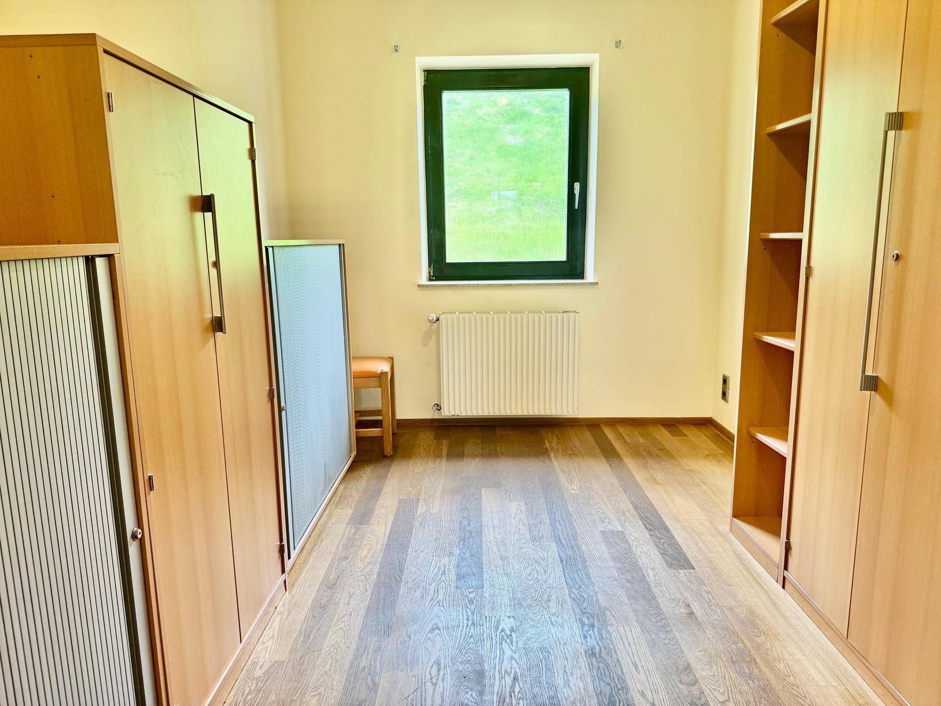 Helles Zimmer mit Holzfußboden, Schränken und einem Fenster mit Blick ins Grüne.