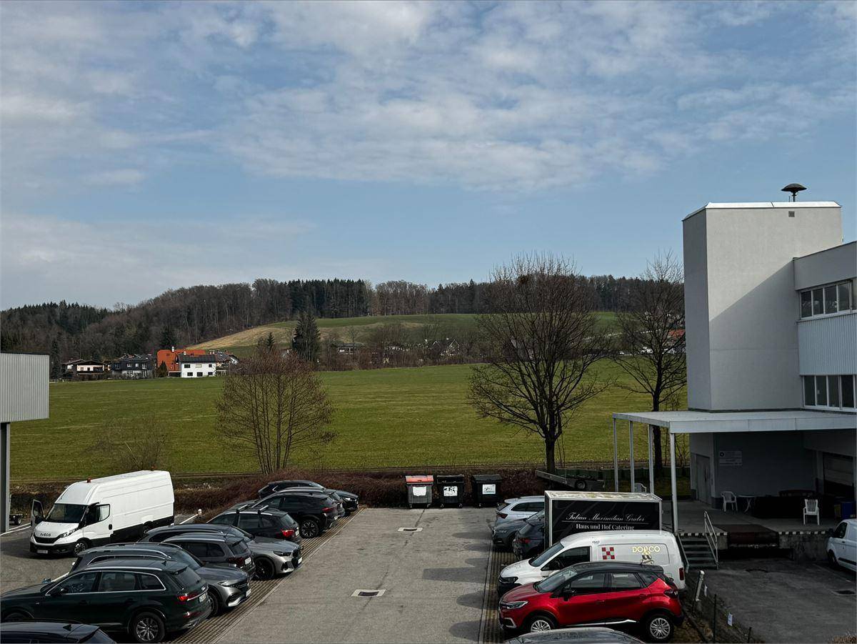 Außenansicht mit Parkplatz, umgeben von grünen Feldern und bewaldeten Hügeln unter blauem Himmel.
