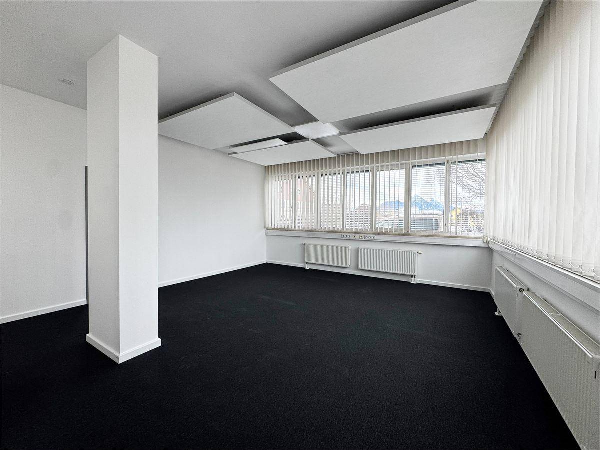 Büroraum mit großen Fenstern, dunklem Teppichboden und akustischen Deckenpaneelen für bessere Raumakustik.