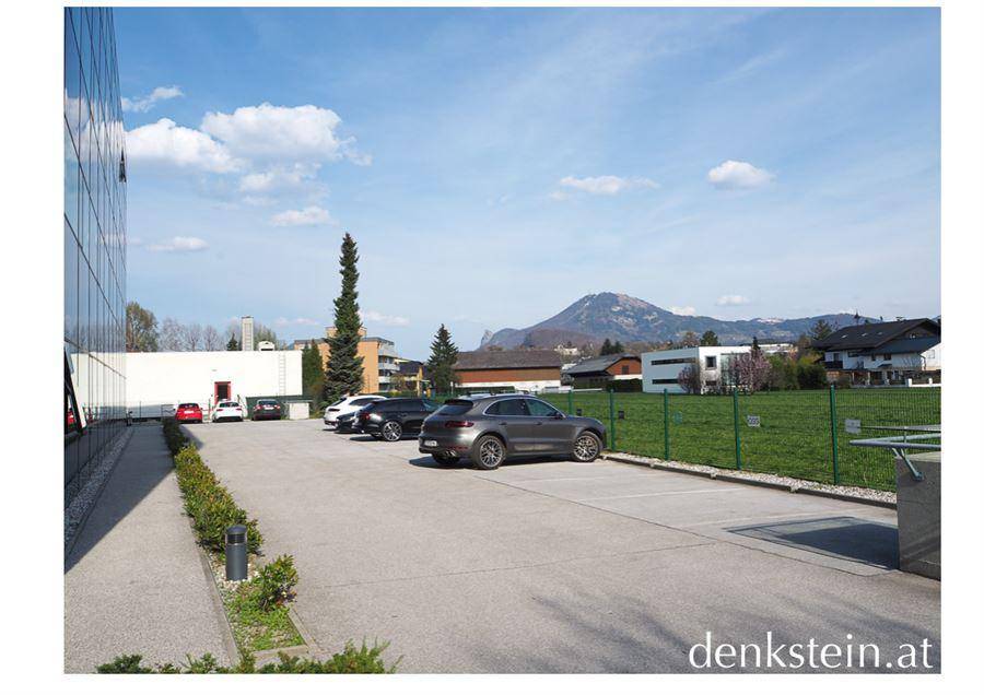 Außenansicht mit Parkplatz, moderner Gebäudefassade und Bergblick im Hintergrund.
