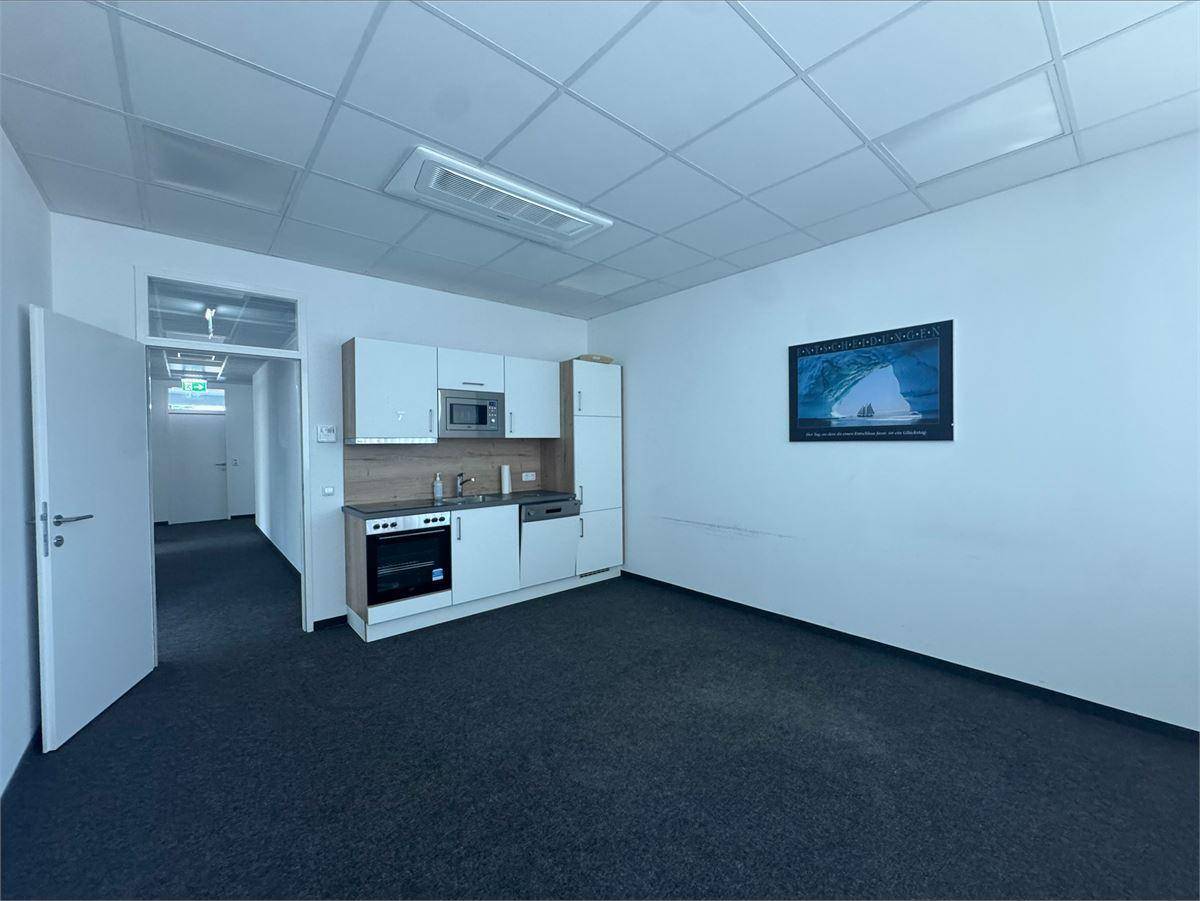 Heller Büroraum mit integrierter Küchenzeile, dunklem Teppichboden und abgehängter Decke.