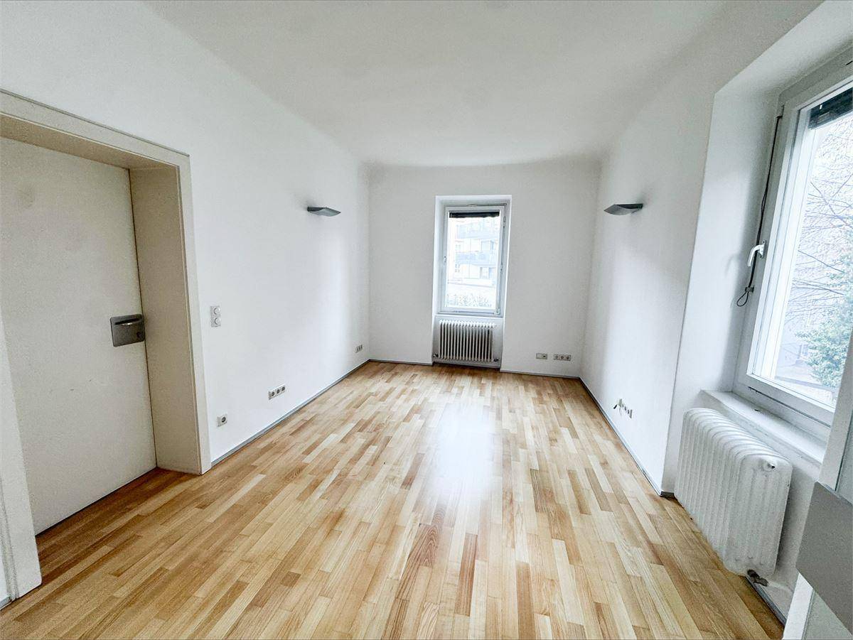 Heller, leerer Raum mit Parkettboden, weißen Wänden und zwei Fenstern mit Heizkörpern darunter.