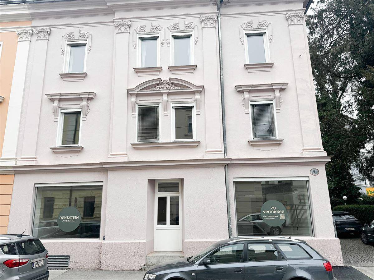 Historisches Gebäude mit aufwendiger Fassade und großen Fenstern im Erdgeschoss, Autos parken davor.